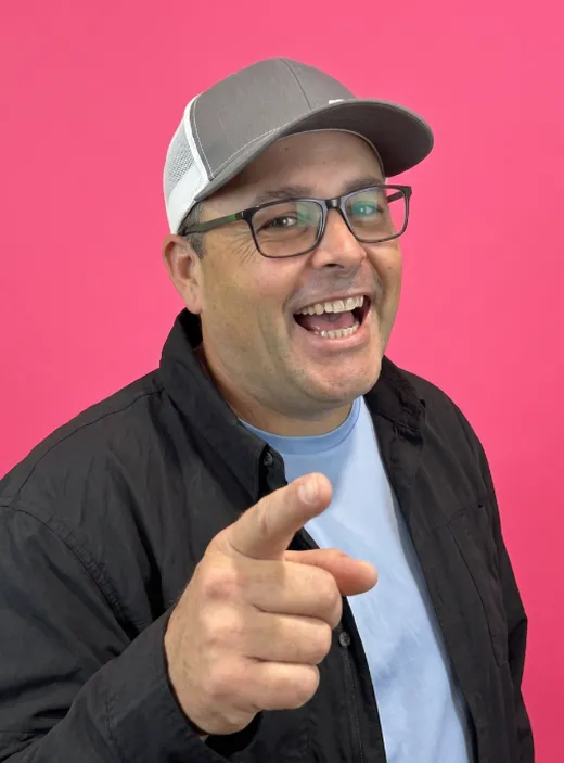 Greg in a trucker cap, laughing and pointing, on a pink background